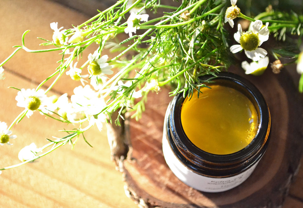 Hand cream jar open and set on a dark wood slice, on a wooden surface, along with chamomile flowers and leaves.