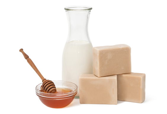 Milk in a glass bottle, honey in glass bowl and wooden dipper, and stacked soap bars.