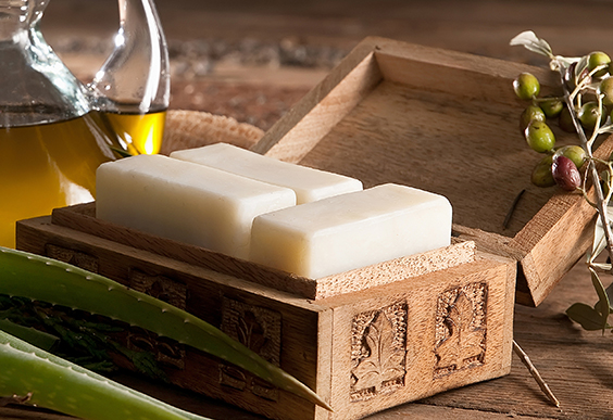 Assorted soap bars in a wooden curved box, aloe vera leaves and olive brach on the sides. Olive oil in a glass jug visible on the back.